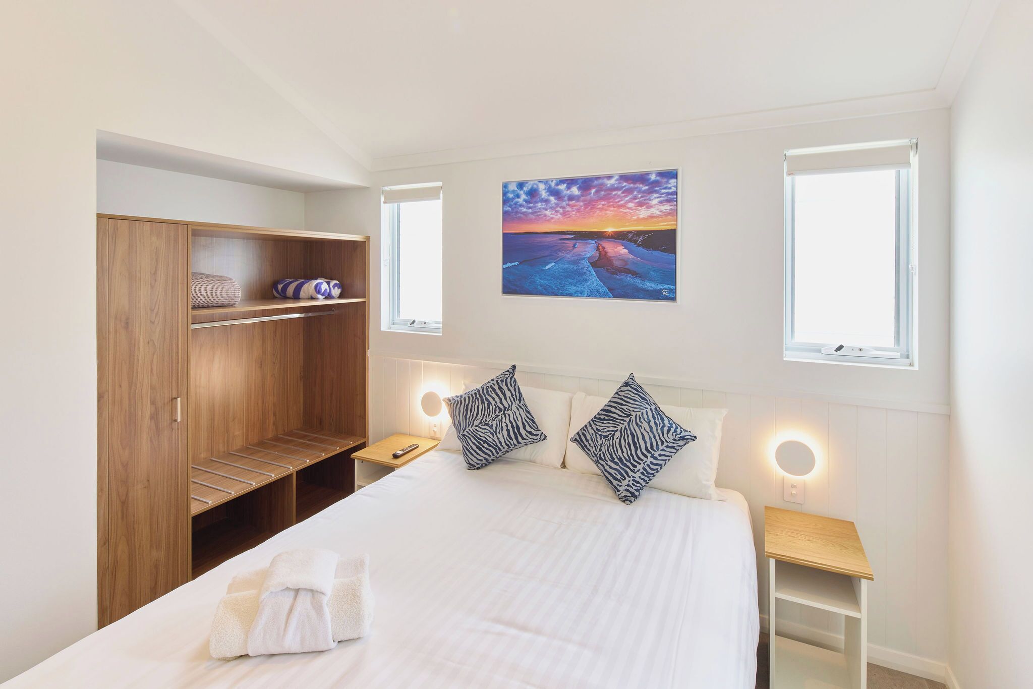 A hotel room with a bed, a wooden wardrobe, a painting of a sunset, and bedside lamps.