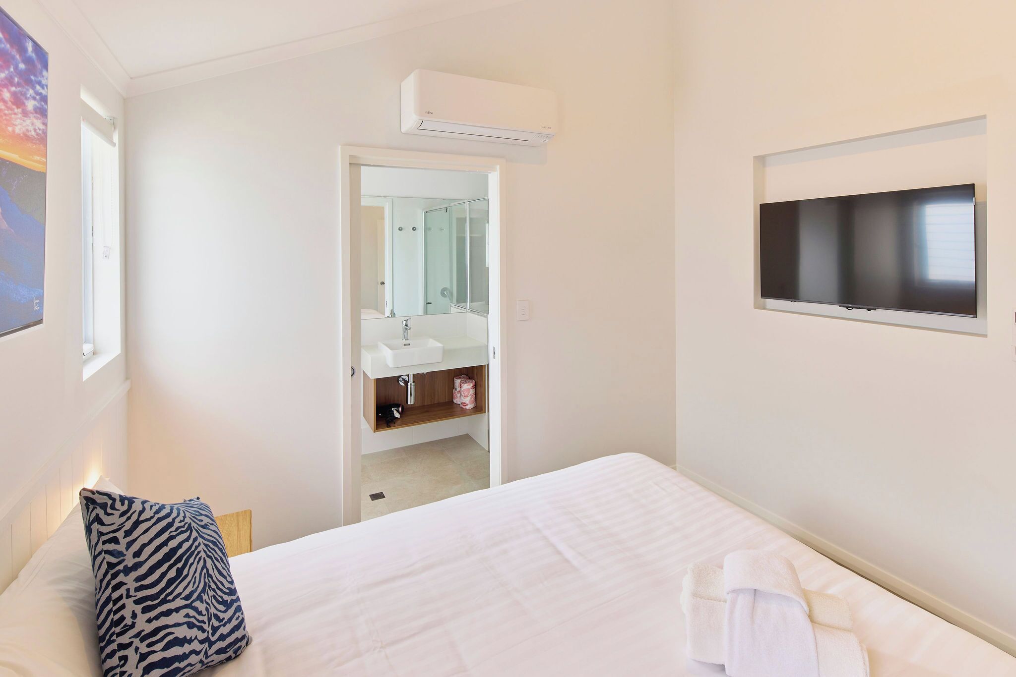 A hotel room with a bed, a TV mounted on the wall, a bathroom with a mirror, and an air conditioning unit.