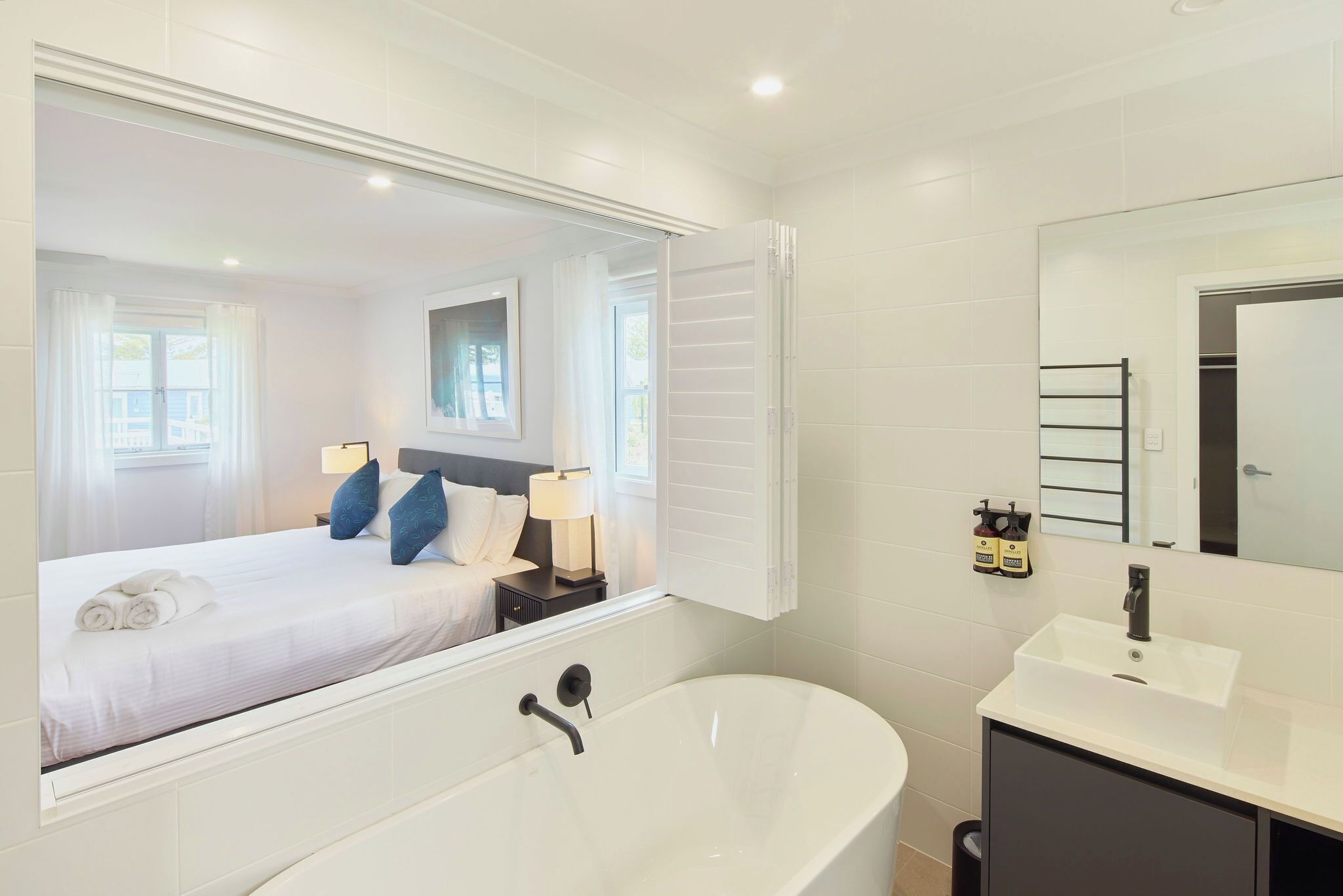A modern bathroom with a large mirror, a bathtub, a sink, and a neatly made bed with pillows and a rolled towel.