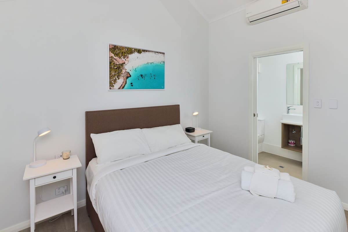 A neatly made bed with white linens, a nightstand with a lamp and a cup, a painting of a beach scene on the wall, and a bathroom visible through an open door.