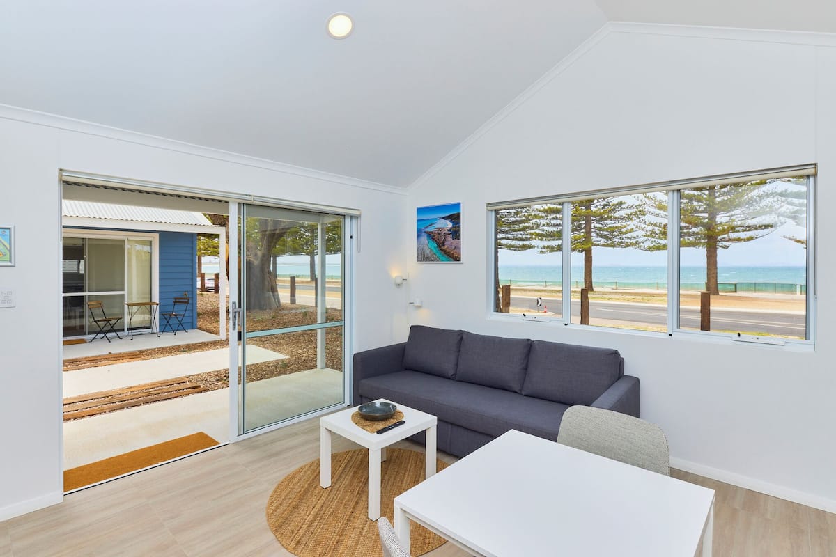 A modern living room with a grey sofa, a white coffee table, and a view of the beach through large windows.
