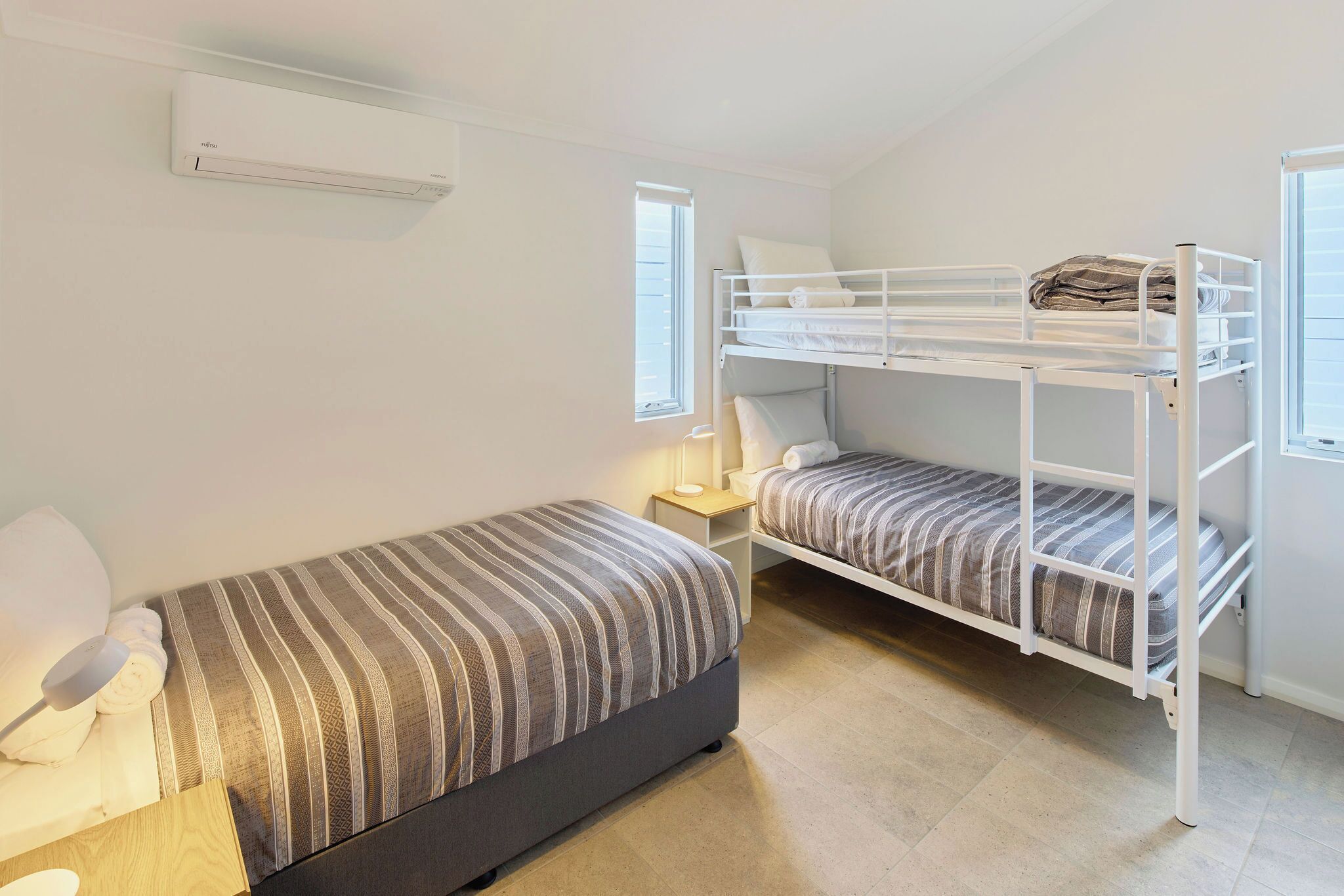 A bedroom with a single bed, a bunk bed, a bedside table with a lamp, and a window.