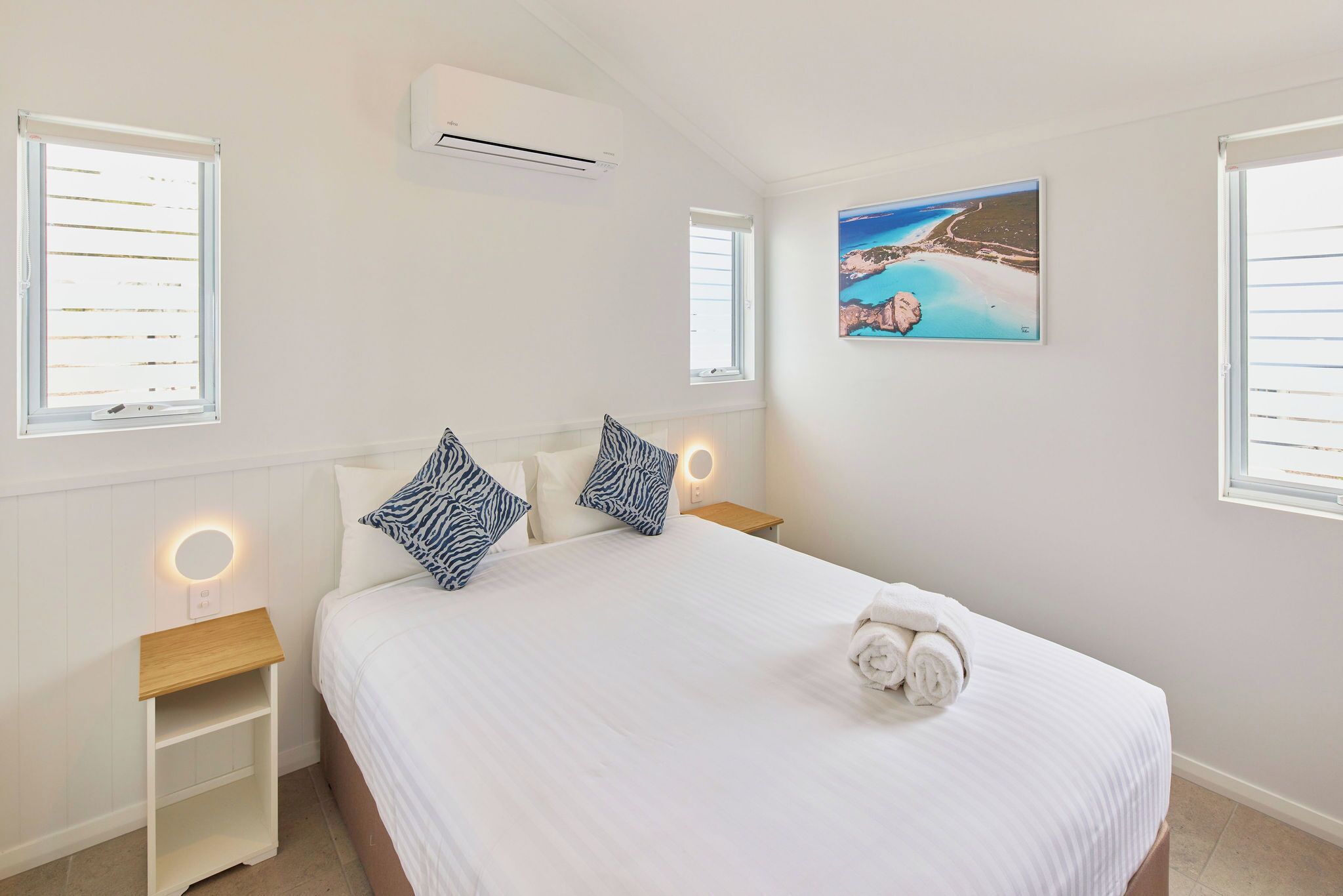 Een slaapkamer met een bed, twee nachtkastjes, een wandairconditioner, een raam met zonwering en een schilderij van een kustlandschap.