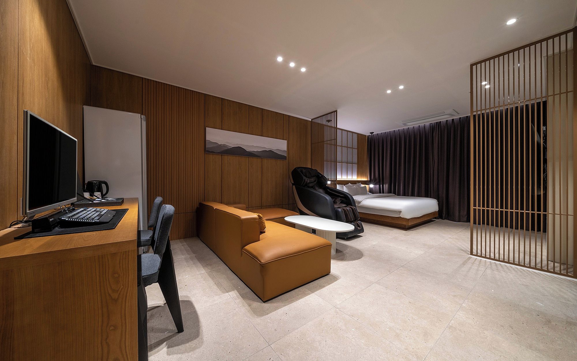 Una habitación de hotel moderna con escritorio, computadora, sillón de masaje, sofá y cama.