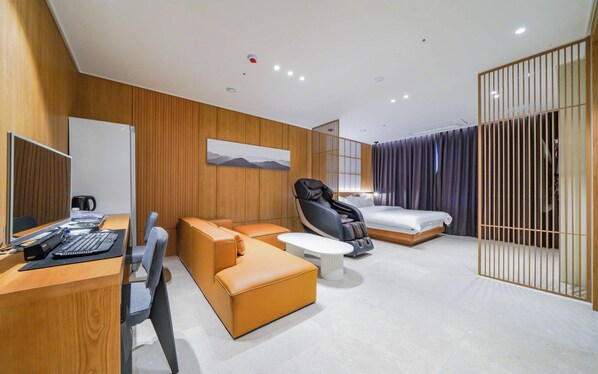 A modern hotel room with a wooden panel wall, a massage chair, a sofa, a bed, and a desk with a computer.
