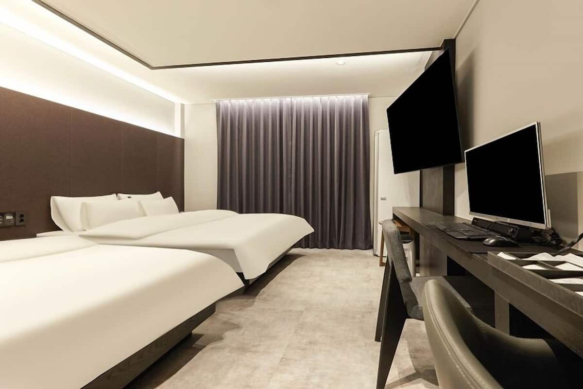 Een moderne hotelkamer met twee bedden, een bureau met een computer en een groot raam met gordijnen.