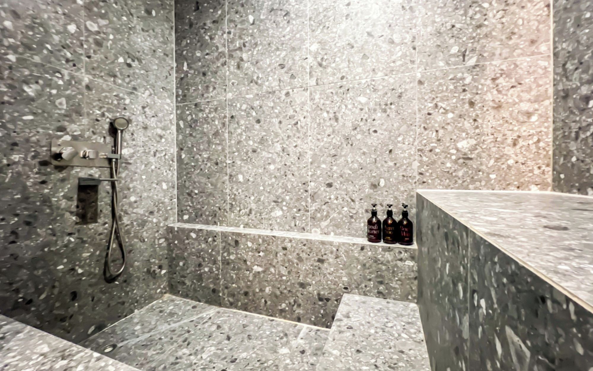 A bathroom with a shower area, a shelf with bottles, and a marble countertop.