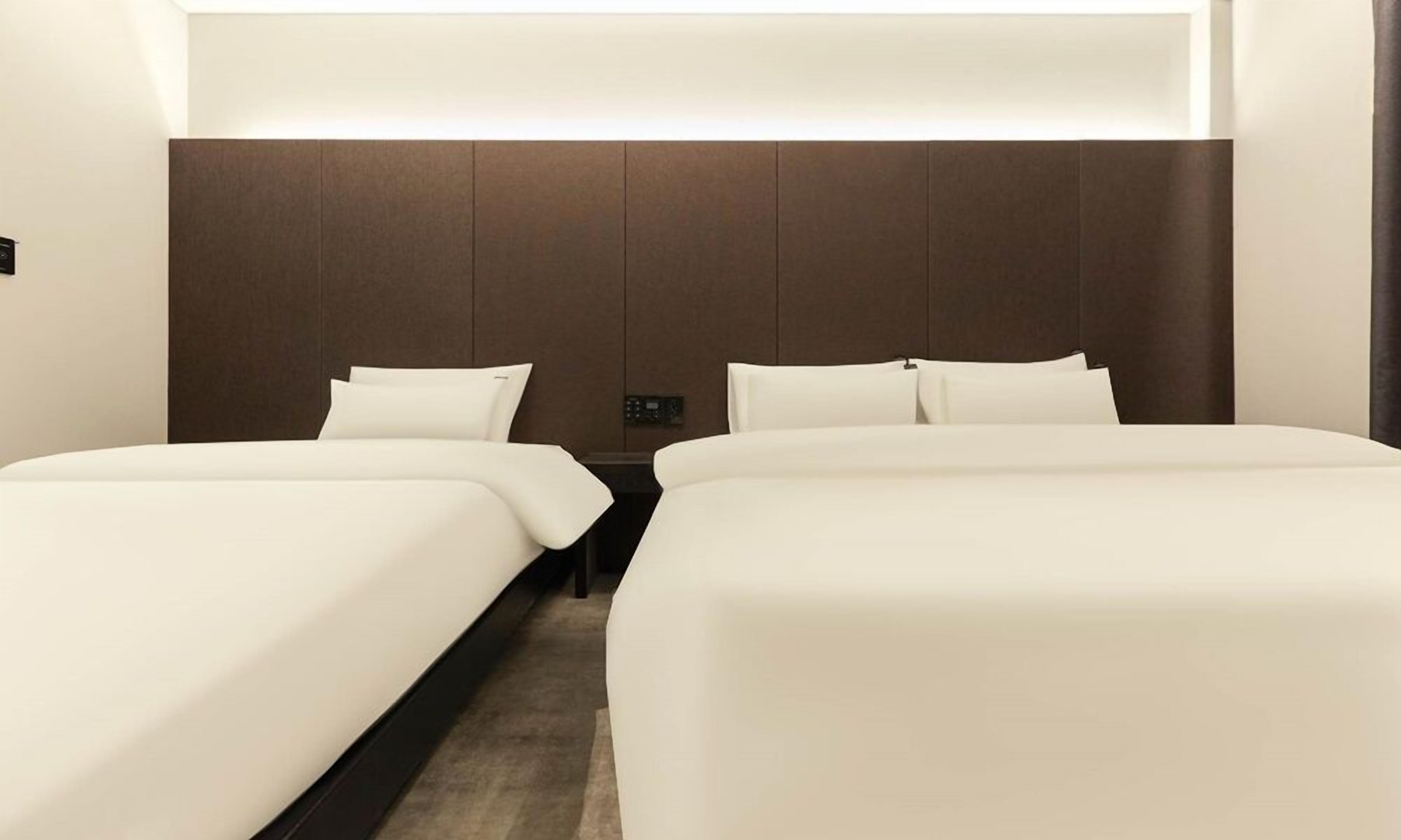 Habitación de hotel con dos camas individuales, un cabecero oscuro y una mesita de noche con panel de control.