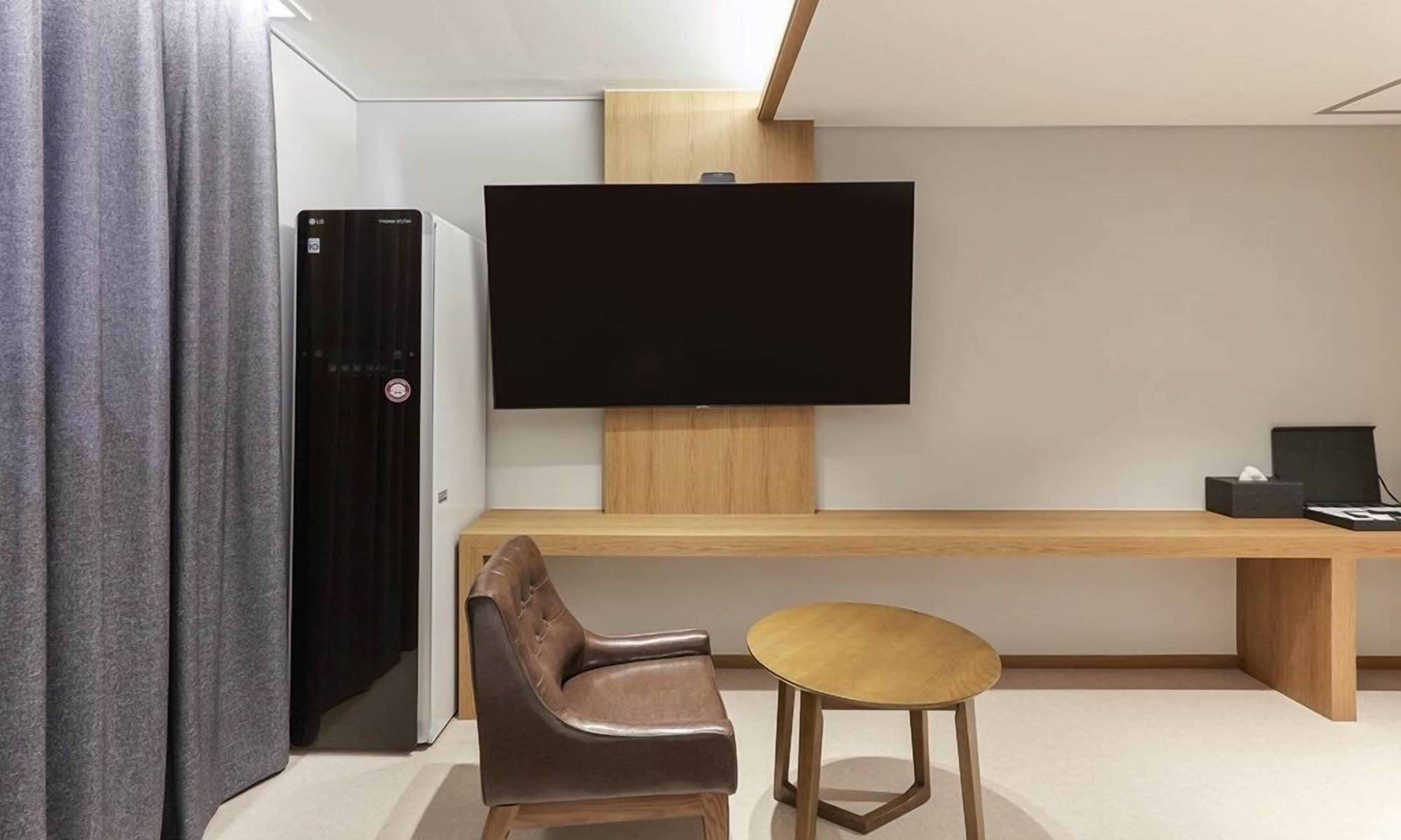 A modern hotel room with a flat-screen TV mounted on the wall, a wooden desk, a leather chair, and a small round table.