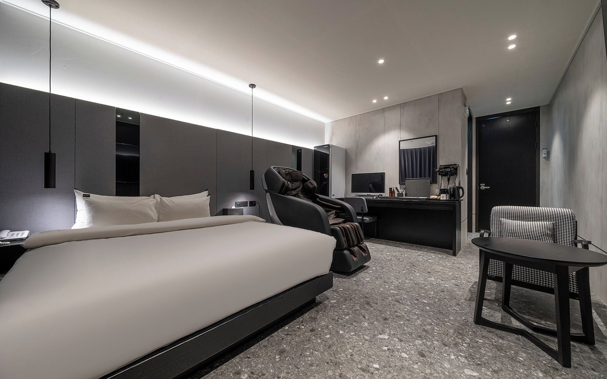 Una habitación de hotel moderna con cama, sillón de masaje, escritorio con computadora y una mesita.