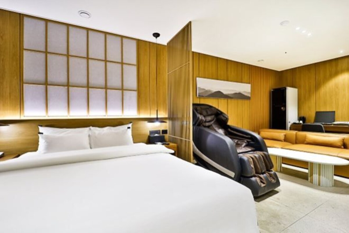 A modern hotel room with a large bed, a massage chair, a sofa, and a desk with a computer.