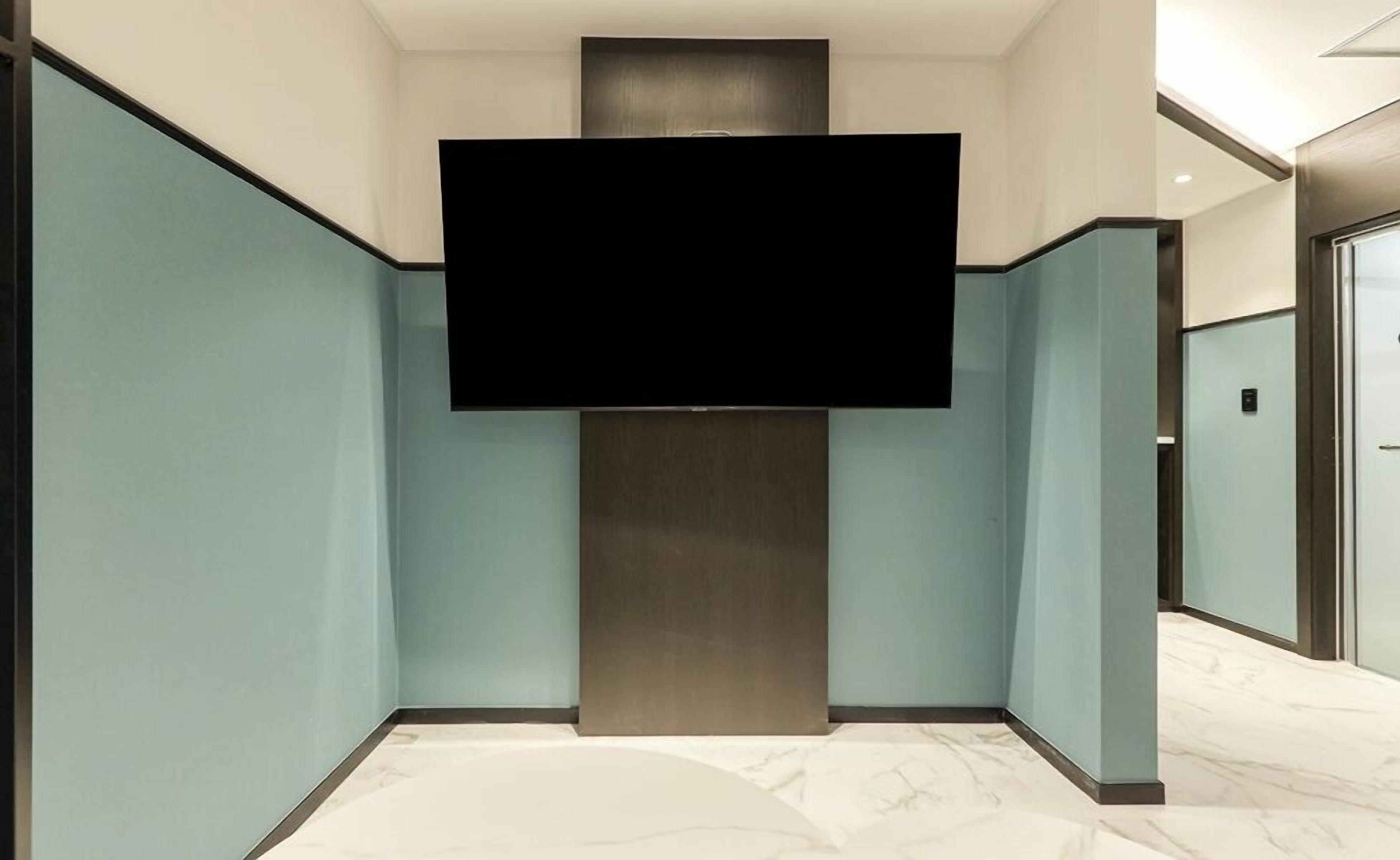 A hallway with light blue walls, a large flat-screen TV mounted on a central column, and a marble floor.