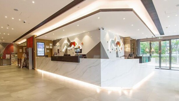 A modern hotel lobby with a reception desk, a digital screen, and a large glass door leading outside.