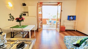 Une pièce compacte comprenant un coin cuisine, un espace repas avec une table et des chaises, un lit recouvert d’une housse à motifs et un téléviseur posé sur un meuble.