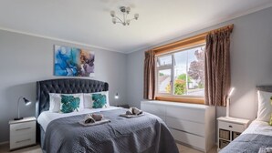 Een slaapkamer met een gestoffeerd hoofdeinde, een groot raam met gordijnen, een nachtkastje met een lamp en een schilderij aan de muur.