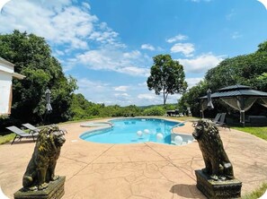 Una piscina con dos estatuas de leones, rodeada por una zona de patio con sillones reclinables y un área de estar cubierta.
