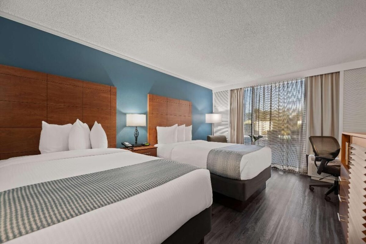 A hotel room with two beds, a desk, a chair, and a large window with curtains.