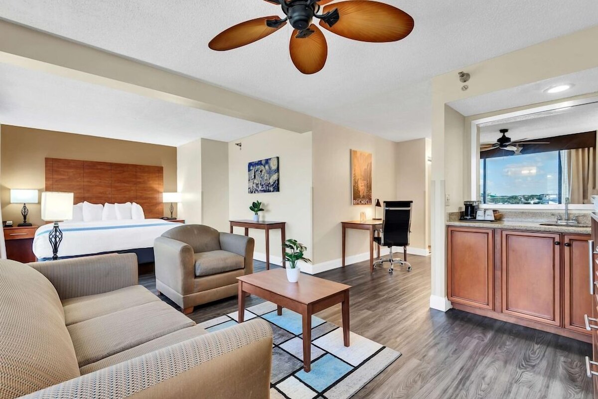 A hotel room with a bed, sofa, coffee table, kitchenette, and a ceiling fan.