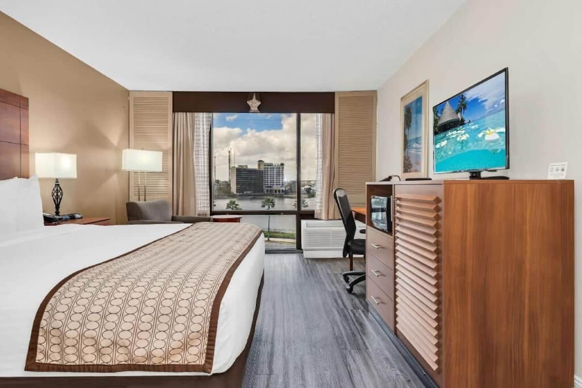A hotel room with a large bed, a desk, a TV, and a view of a cityscape through large windows.