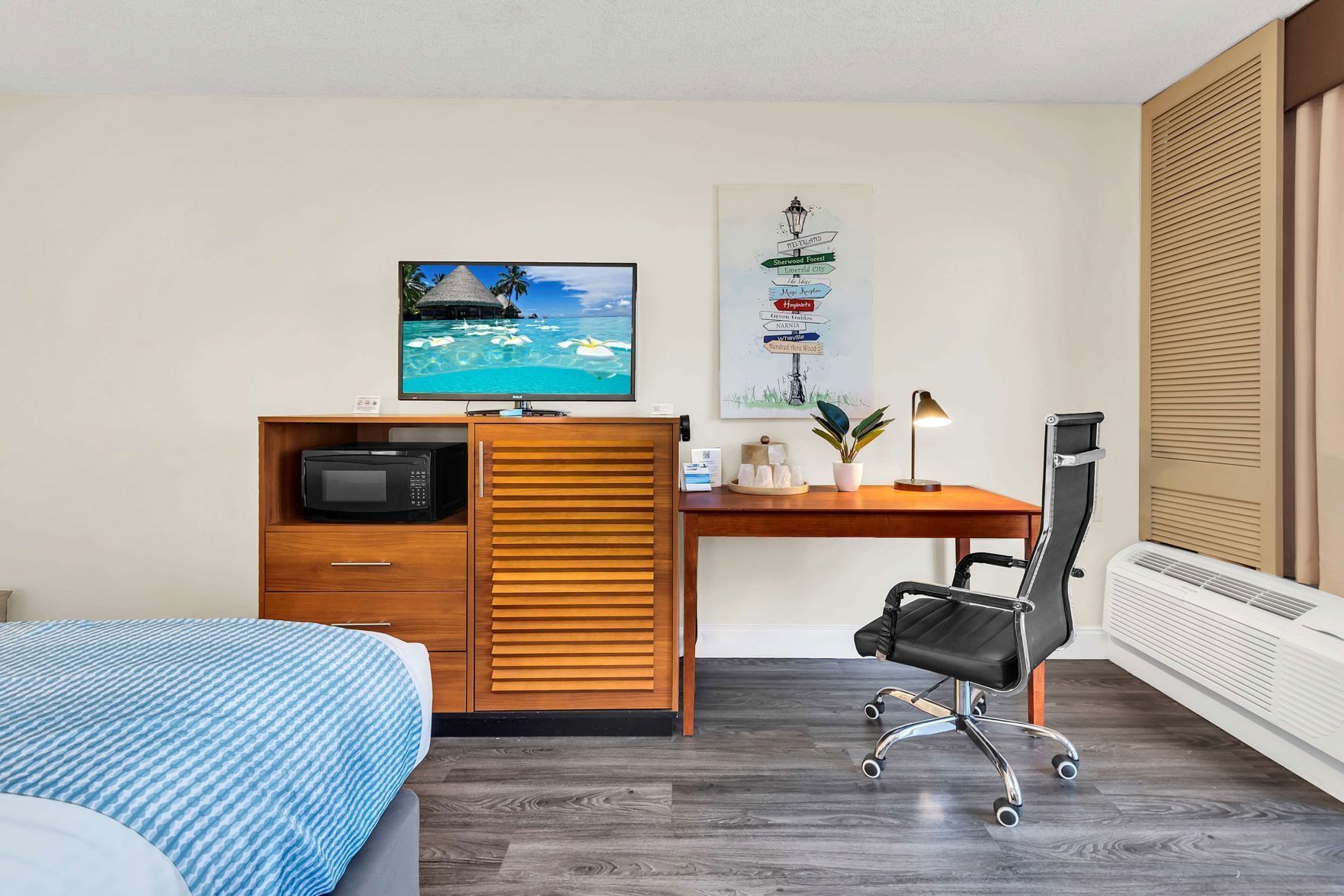 A hotel room with a bed, a desk with a computer, a chair, a TV, and a poster on the wall.