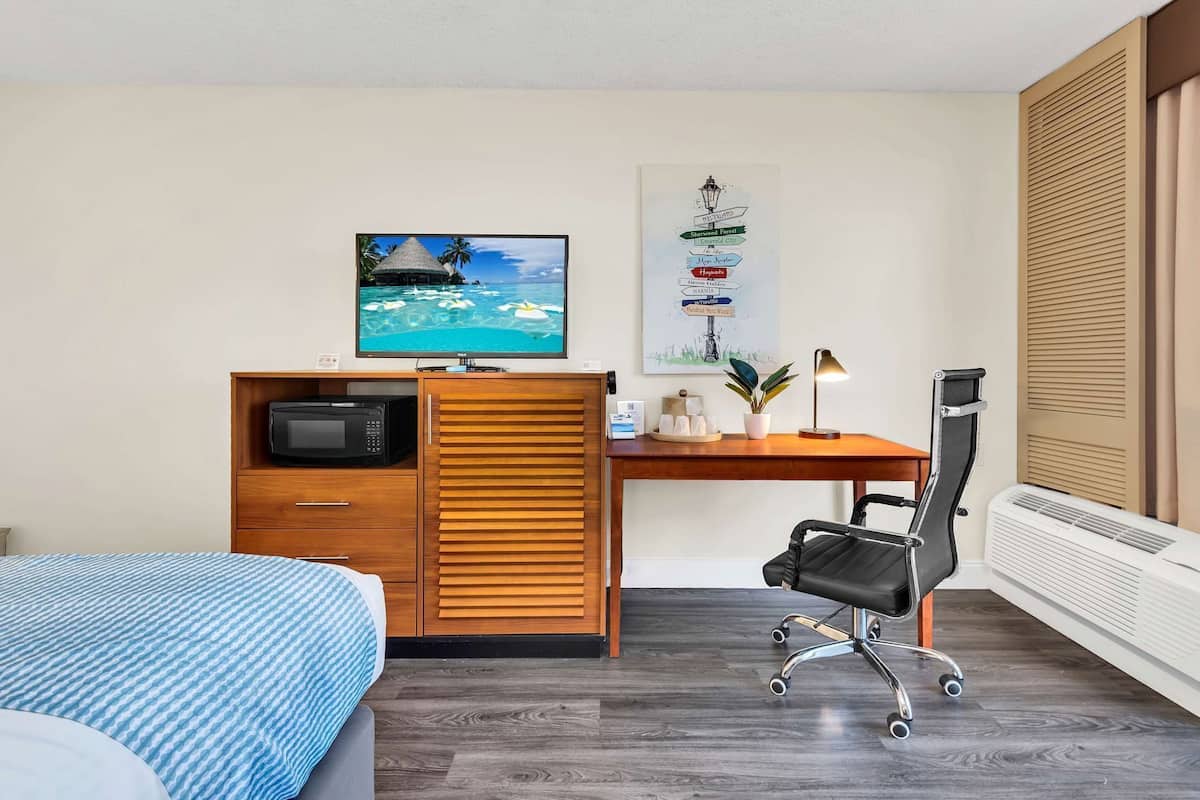 A hotel room with a bed, a desk with a computer, a chair, a TV, and a poster on the wall.