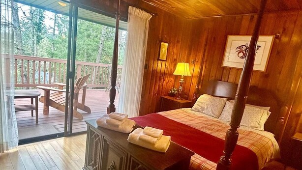 Un lit à baldaquin avec une couvre-lit à carreaux rouges et blancs, une tête de lit en bois et une table de chevet avec une lampe. Sur un balcon donnant sur les arbres, on trouve un bureau et une chaise en bois.