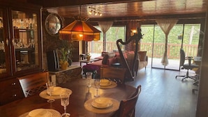 Un coin repas avec une table en bois dressée pour quatre personnes, entourée de chaises. On aperçoit une harpe au fond, et une grande fenêtre avec des rideaux.
