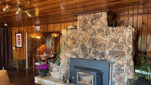 Une cheminée en pierre avec un encadrement noir, un plafond en bois et un coin repas comprenant une table et des chaises.