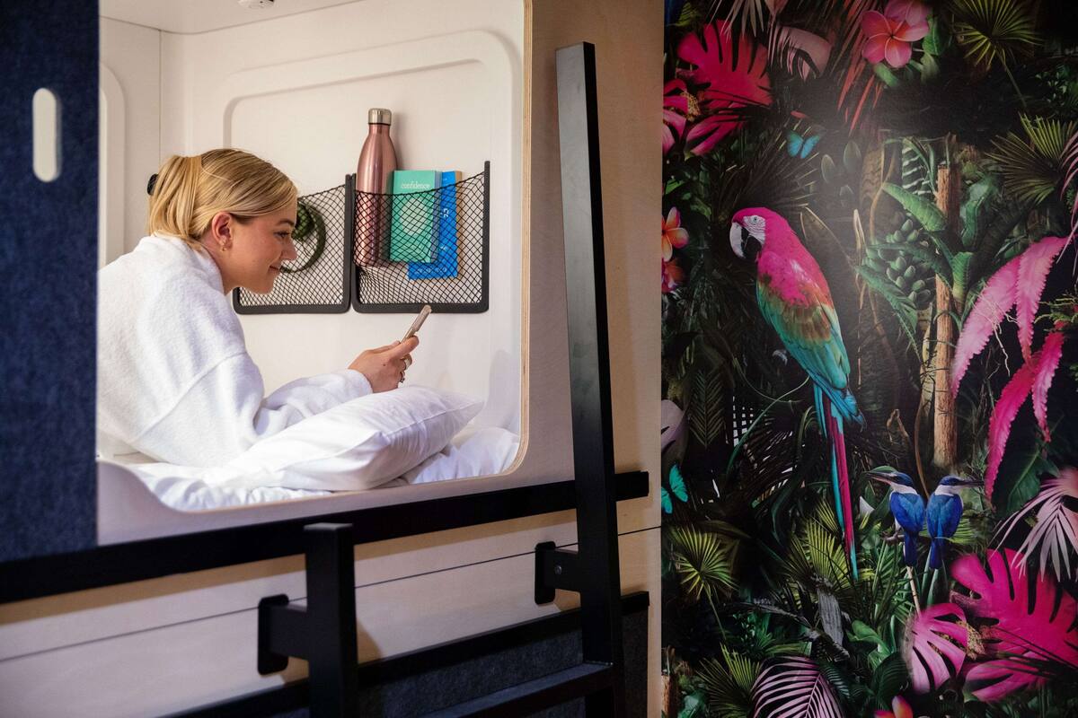 A person in a white robe sitting in a room with a bed, a window, and a wall with a tropical mural.