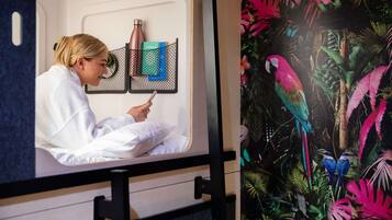 Una persona vestida con una bata blanca sentada en una habitación con una cama, una ventana y un mural tropical en la pared.