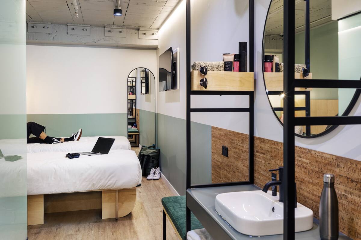A modern hotel room with a bed, a desk, a mirror, and a bathroom with a sink and mirror.