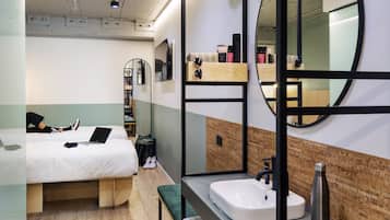 A modern hotel room with a bed, a desk, a mirror, and a bathroom with a sink and mirror.