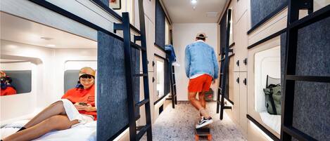 A narrow corridor with bunk beds on either side, a person skateboarding, and another person sitting on a bed.