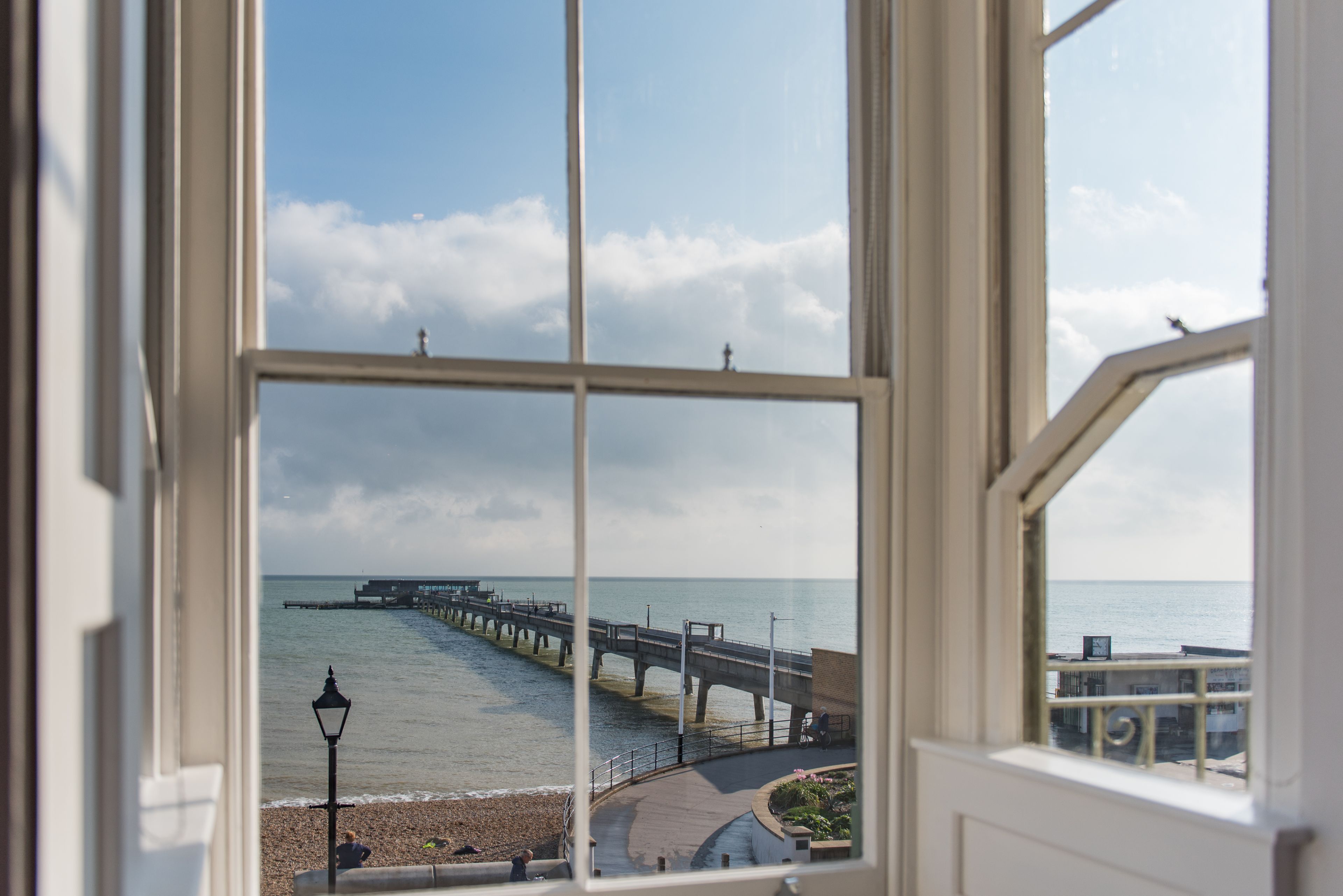 Une vue à travers une fenêtre sur un paysage côtier, avec une jetée s’avançant dans l’eau, une plage et un bâtiment doté d’un balcon.