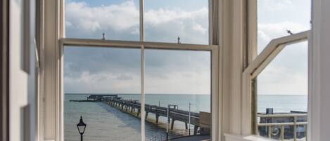 Une vue à travers une fenêtre sur un paysage côtier, avec une jetée s’avançant dans l’eau, une plage et un bâtiment doté d’un balcon.