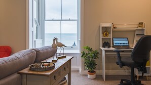 A living room with a view of the sea, a desk with a laptop, a red chair, and a wooden coffee table with decorative items. - Light, bright, and colourful apartment opposite Deal pier (Deal)