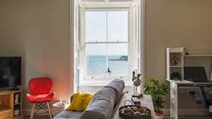 A living room with a large window, a red chair, a grey sofa, a wooden table, and a TV. - Light, bright, and colourful apartment opposite Deal pier (Deal)