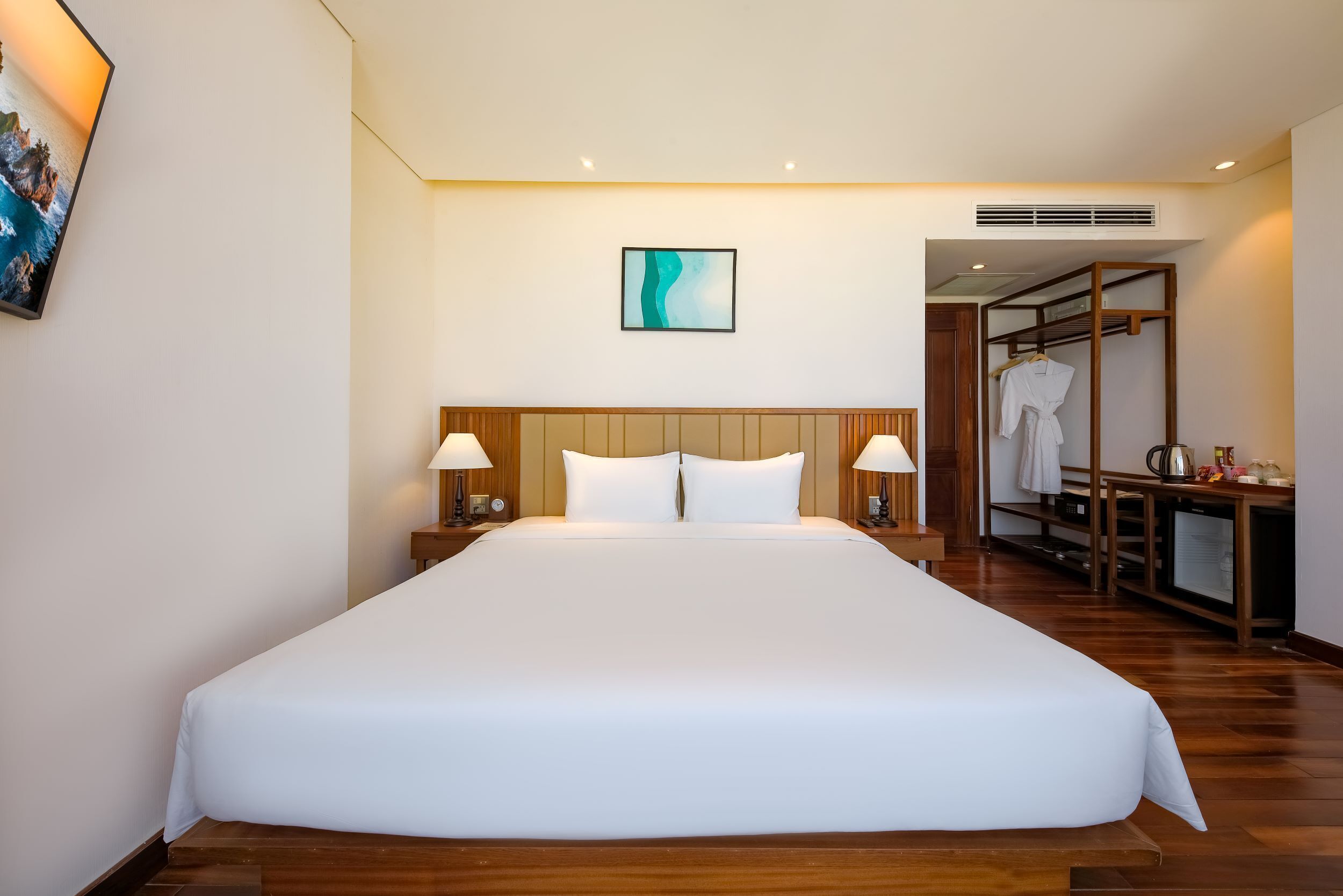 Uma cama grande com roupa de cama branca, cabeceira de madeira, duas luminárias de apoio e uma pintura abstrata emoldurada na parede.