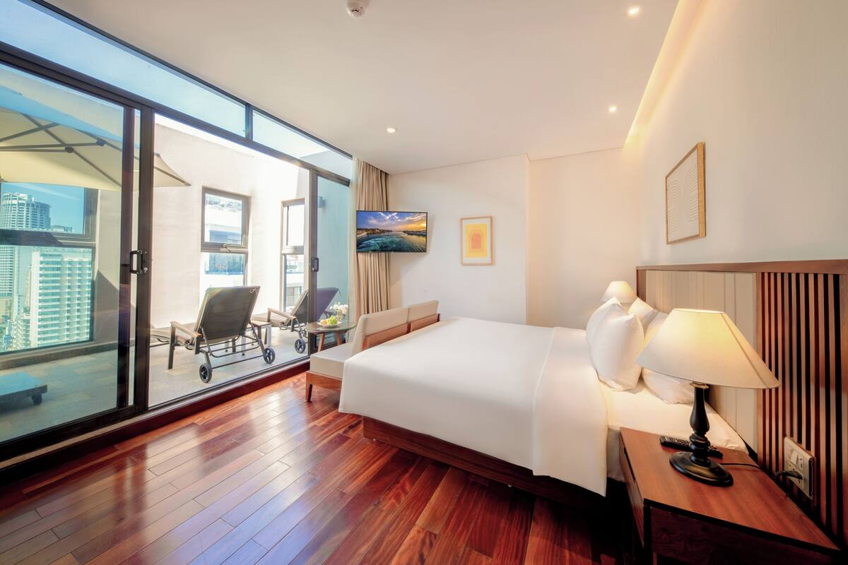 A hotel room with a large bed, a desk with a lamp, a chair, a TV, and a sliding glass door leading to a balcony with a view of buildings.