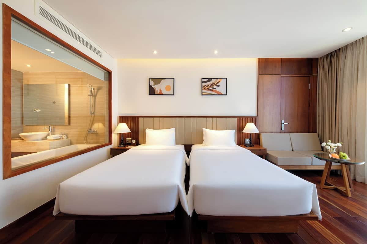 A hotel room with two single beds, a large mirror, a shower, a small sofa, and two wall-mounted paintings.
