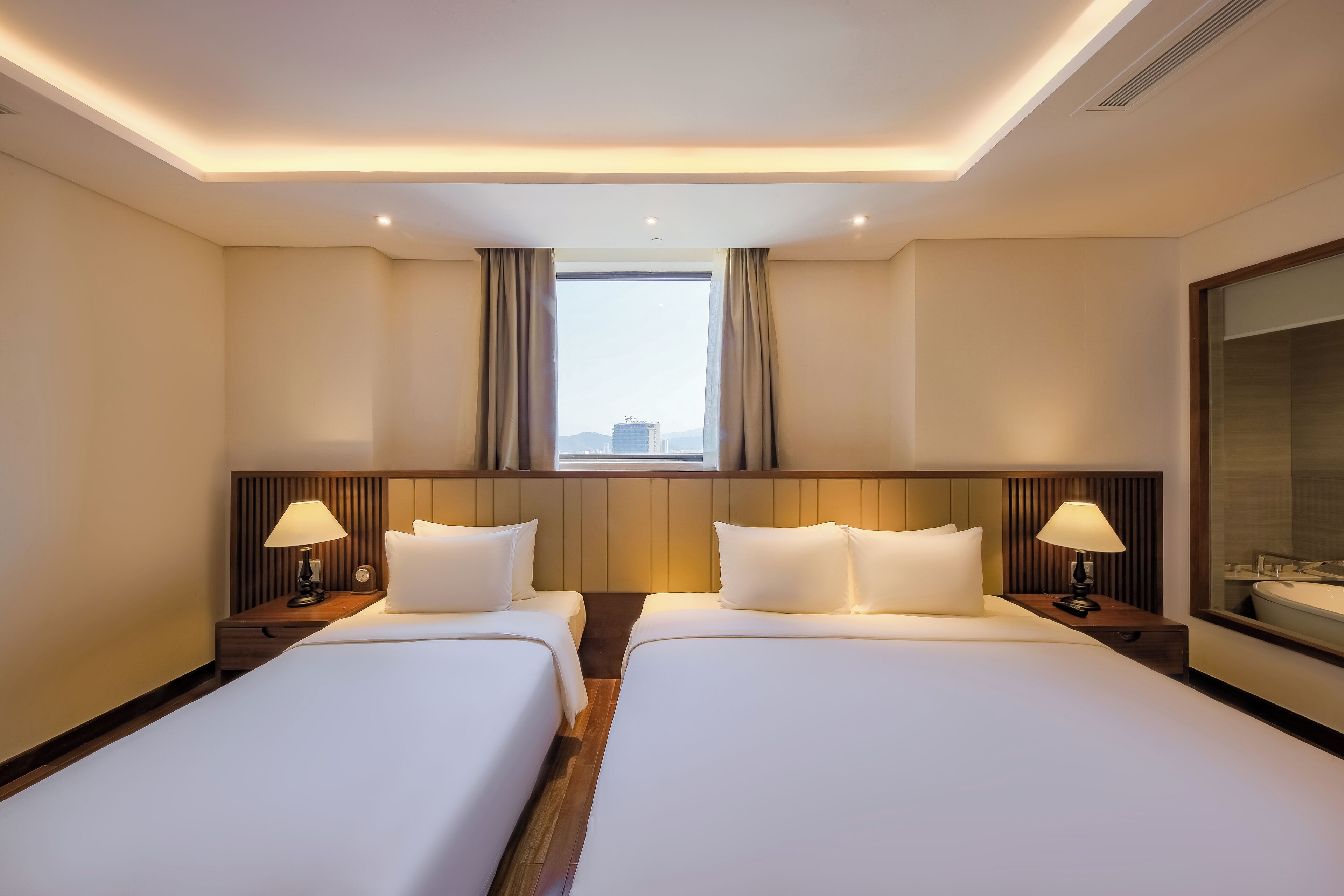 A hotel room with two beds, a large window with curtains, a nightstand with a lamp, and a bathroom with a mirror.
