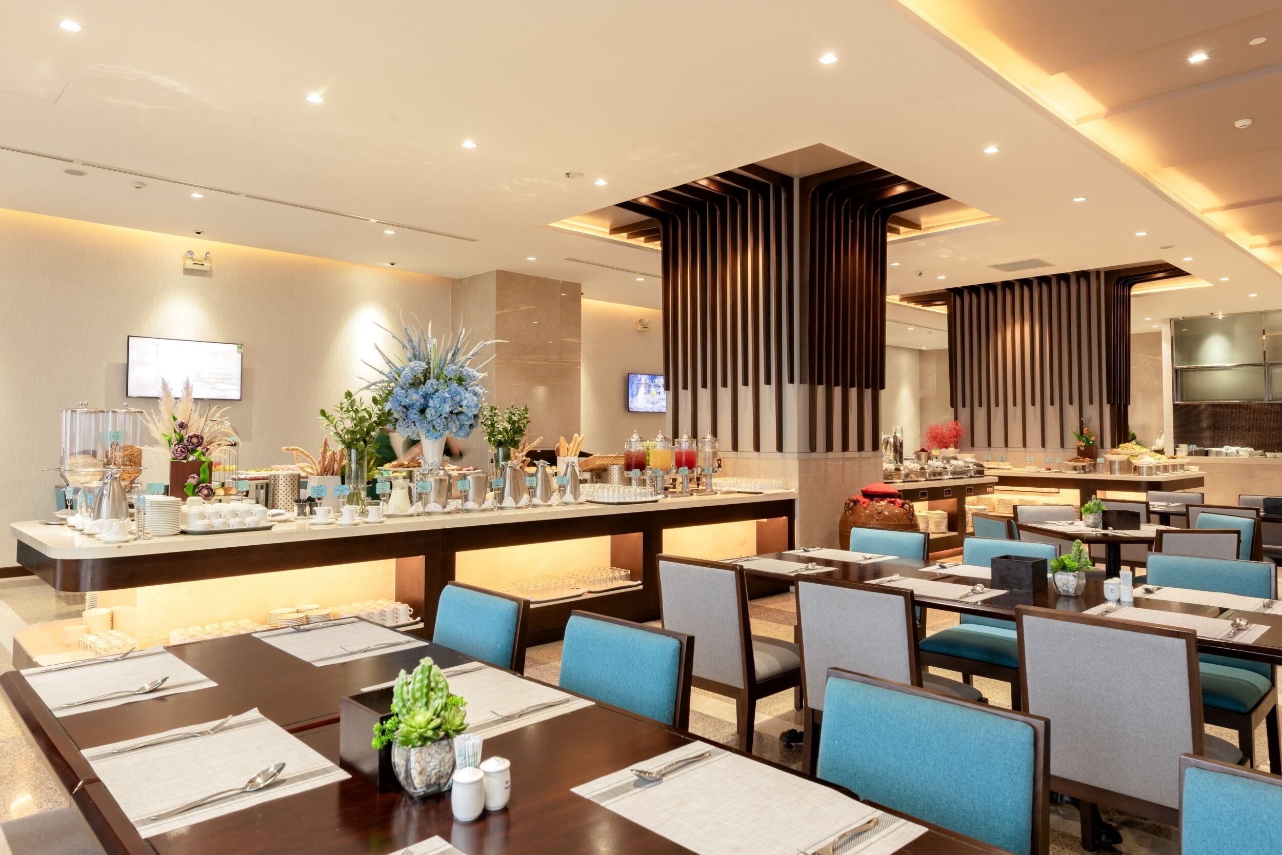 A modern hotel dining area with a buffet setup, multiple dining tables, and a central island with a variety of food and drink options.