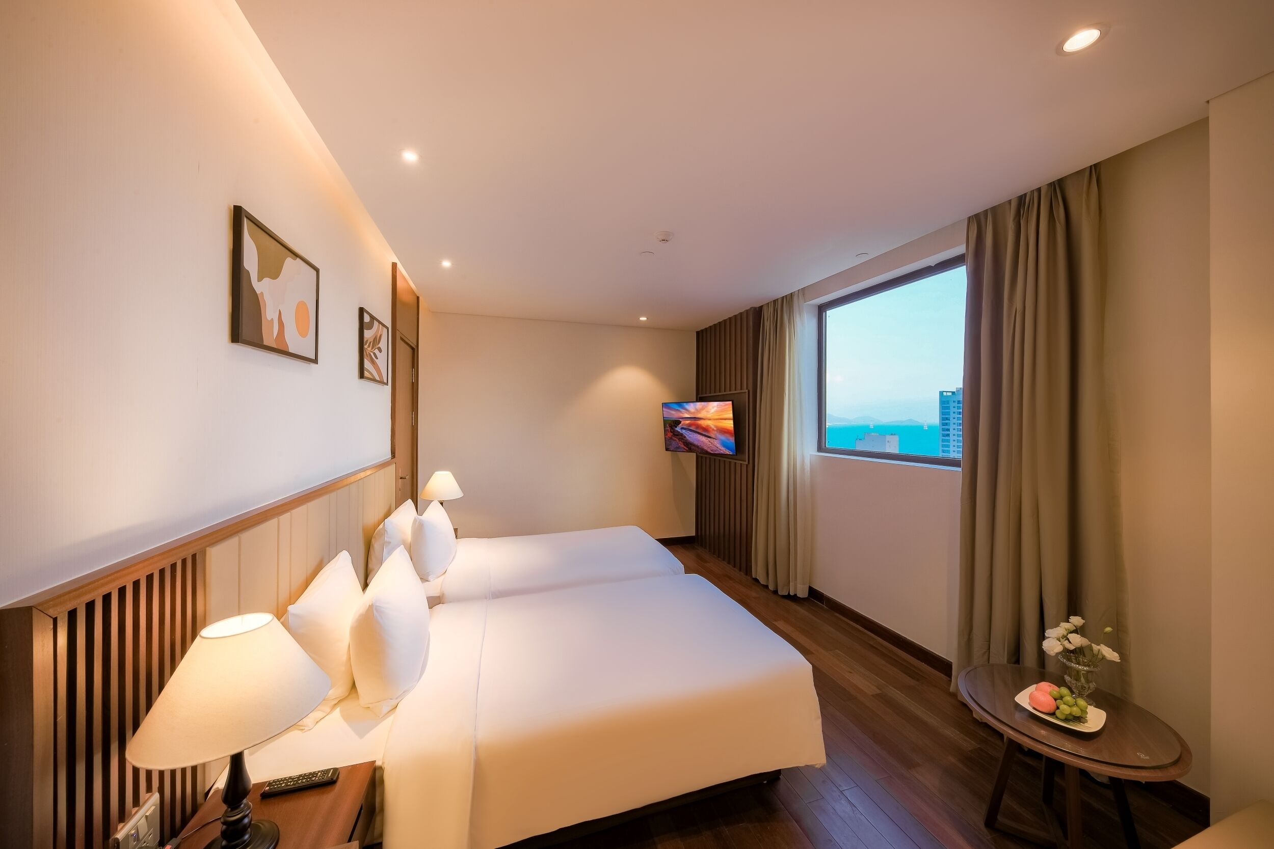 A hotel room with a large bed, a TV, a small table with flowers, and a view of the city.