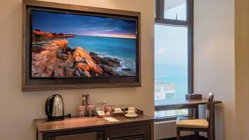 A room with a large flat-screen TV mounted on the wall, a wooden cabinet below, and a window offering a view of the sea.