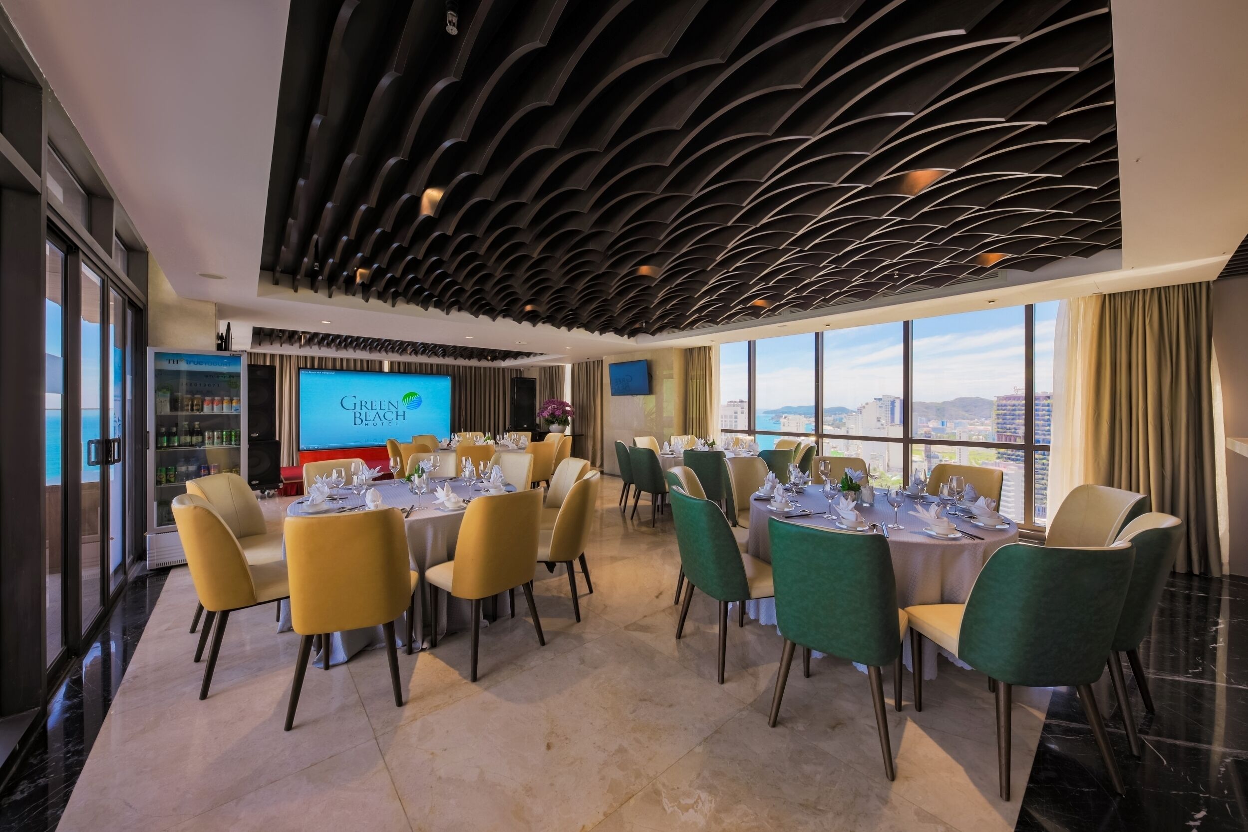 Un área de comedor moderna con mesas redondas, sillas amarillas y verdes, y una pantalla grande que muestra 'Green Beach'.