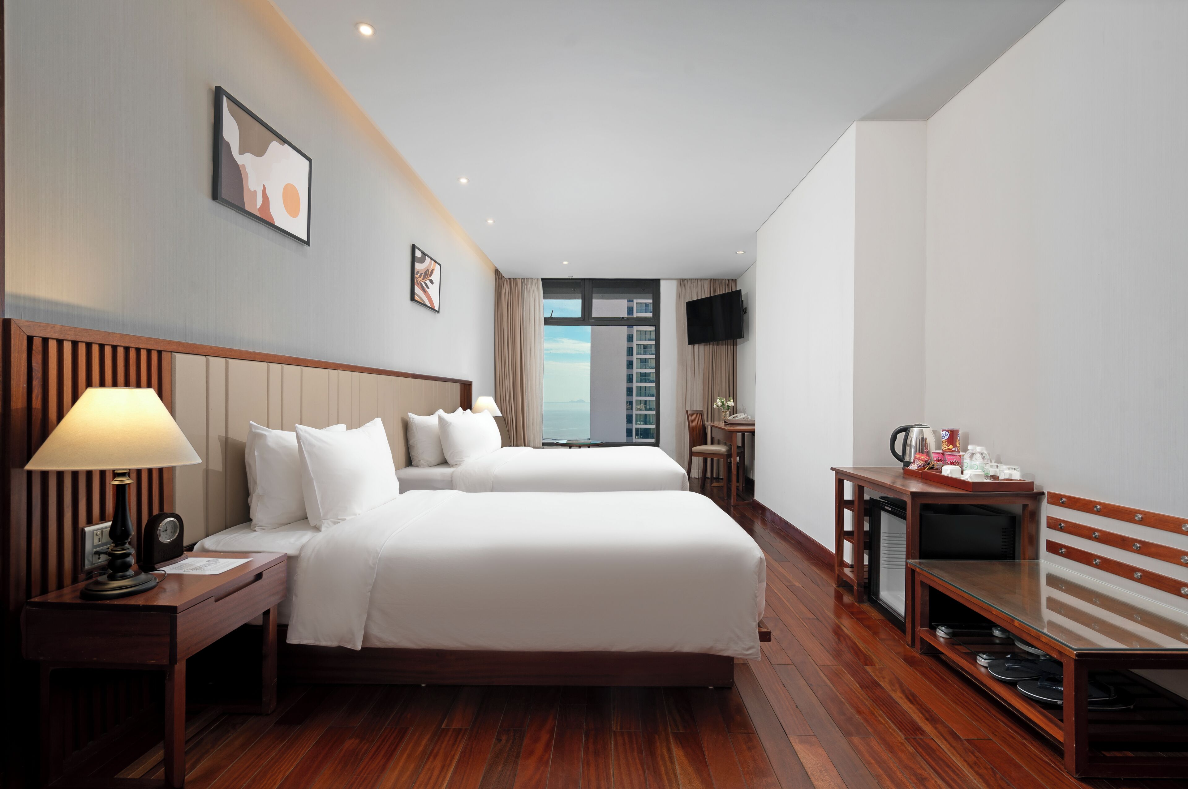 Habitación de hotel con dos camas, un escritorio con lámpara, televisión y vistas a la ciudad.