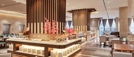 A modern hotel lobby with a central buffet area, seating arrangements, and a large window providing a view of the city.