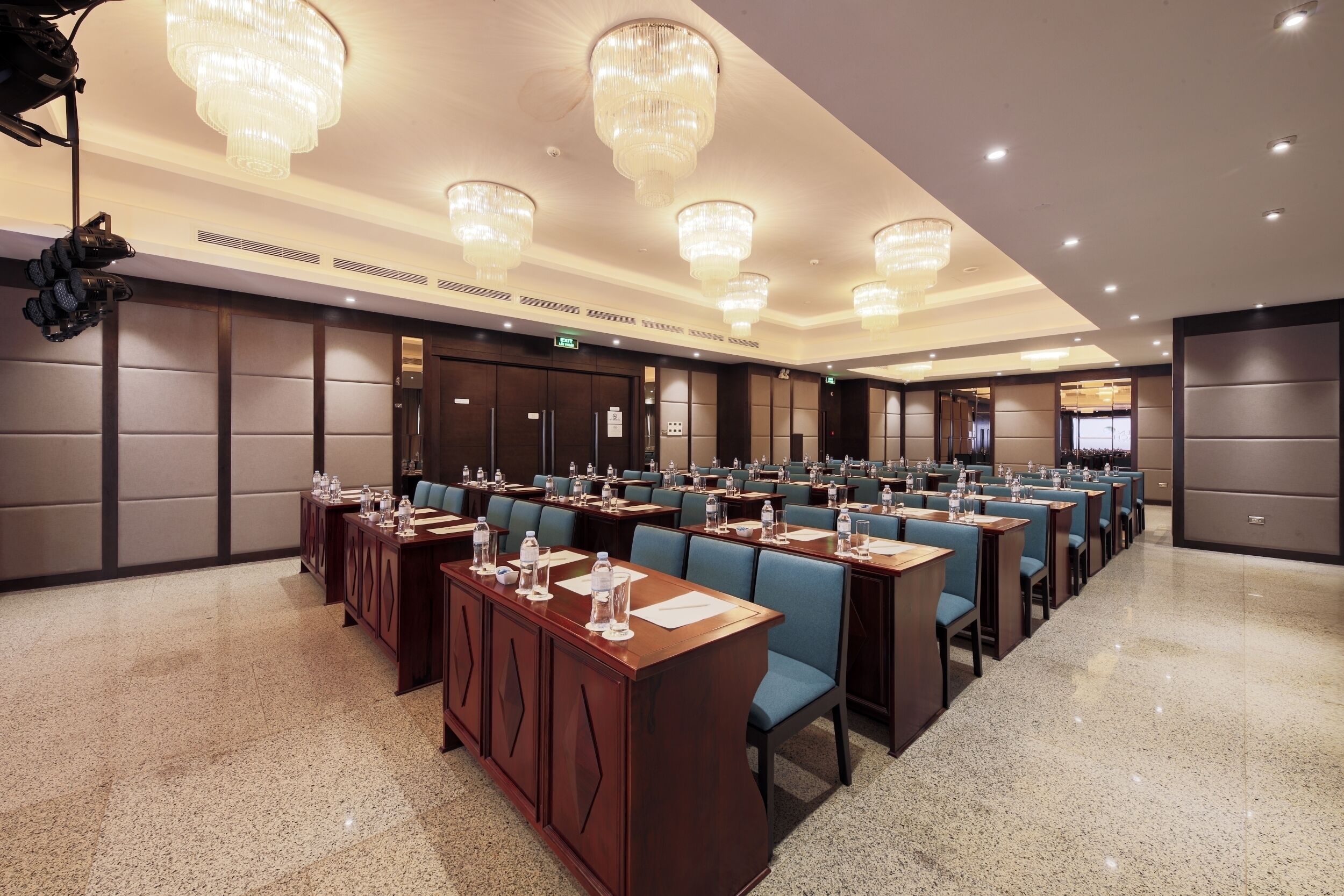 A conference room with rows of tables and chairs, large chandeliers, and a polished floor.