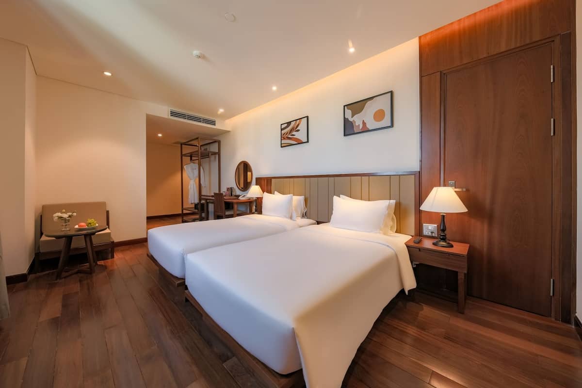 A hotel room with two beds, a desk, a chair, a lamp, and artwork on the walls.