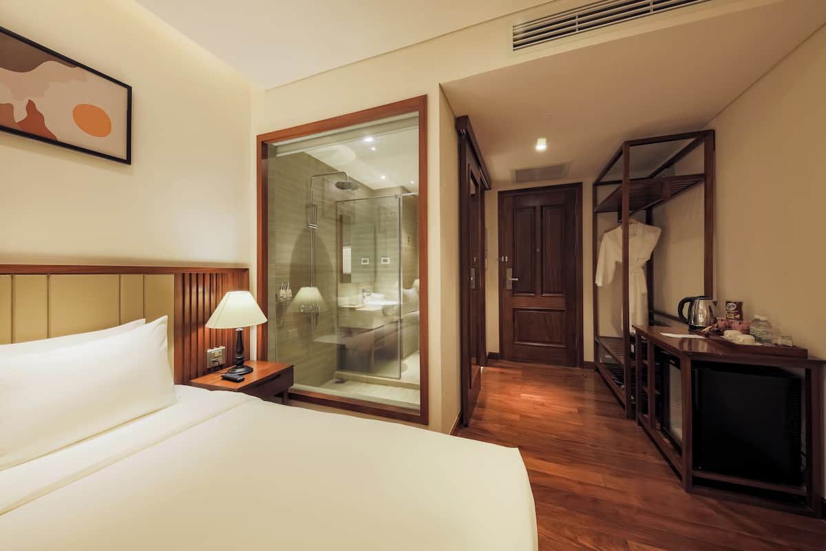 A hotel room with a bed, bedside lamp, a glass-enclosed shower, a wooden wardrobe, and a mirror.